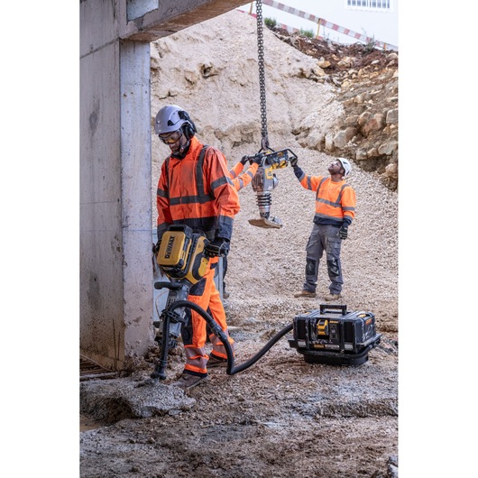 Workers in orange gear use DEWALT tools at a dusty construction site with heavy machinery and a portable power unit.