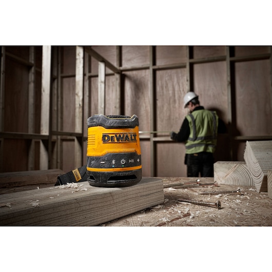 A DEWALT DCR009 portable Bluetooth speaker placed on a wooden plank in a construction site, with a worker in a safety vest and helmet measuring in the background.