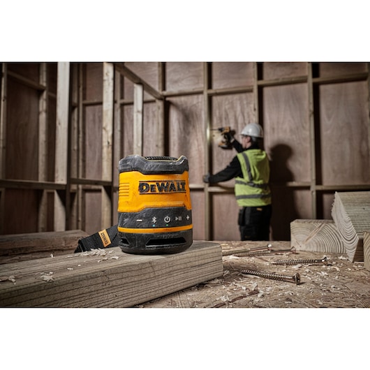 A DEWALT DCR009 portable speaker placed on a wooden beam at a construction site, with a worker in safety gear operating a tool in the background.