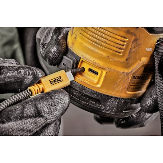 Close-up of a person wearing black gloves plugging a yellow DEWALT USB-C charging cable into a DEWALT DCR009 device. The device has visible scratches and a product label.