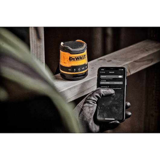 A DEWALT DCR009 portable Bluetooth speaker sits on a wooden surface, while a person wearing a work glove operates a smartphone showing the speaker connected via Bluetooth.