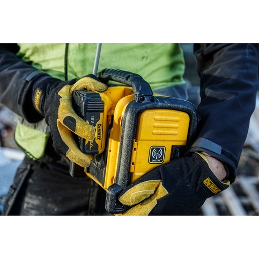 A person wearing DEWALT branded work gloves is holding a yellow DEWALT DCR020 portable digital radio outdoors. The radio is