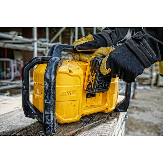 Gloved hands insert battery into yellow DEWALT device at construction site with scaffolding in background.