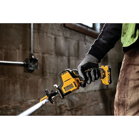 Gloved hand holding a DEWALT DCS312D2 cordless reciprocating saw cutting a metal pipe indoors.