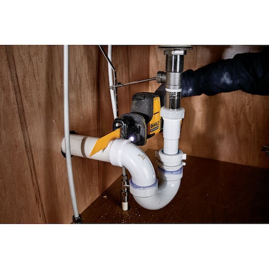DEWALT cordless saw cutting a PVC pipe under a sink in a wooden cabinet.