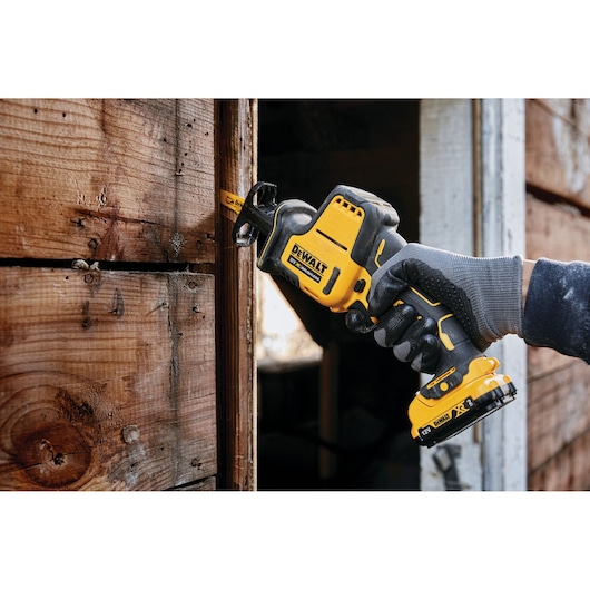 Hand using a DEWALT cordless reciprocating saw to cut wooden boards near a window frame outside a building.