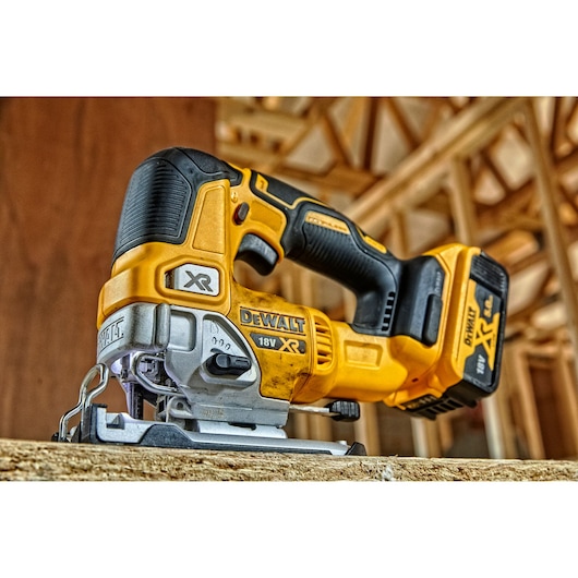 A close-up of a DEWALT DCS334N cordless jigsaw, placed on a wooden surface in a construction environment. The tool is yellow and black, and features a visible battery labeled 18V XR.