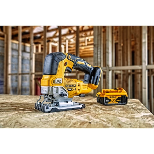 A DEWALT 18V cordless jigsaw and a DEWALT XR 5.0Ah battery are placed on a wooden surface inside a construction site with unfinished wooden framing.