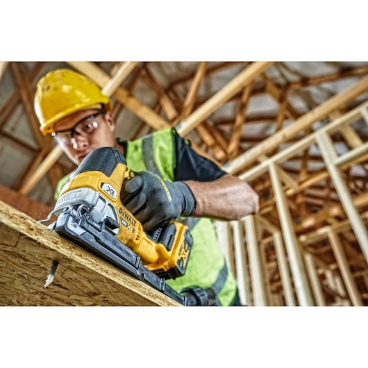 A person wearing a safety helmet and high-visibility vest uses a DEWALT cordless jigsaw (model DCS334P2) to cut wood at a construction site with exposed timber beams.