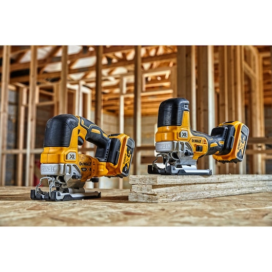 Two DEWALT cordless jigsaws are placed on a wooden surface in a construction site with unfinished wooden framing in the background.
