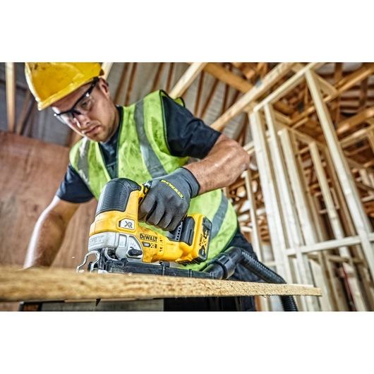 A person wearing a yellow hard hat, safety vest, and gloves is using a DEWALT DCS334P2 cordless jigsaw to cut a wooden board at a construction site with unfinished wooden framing.