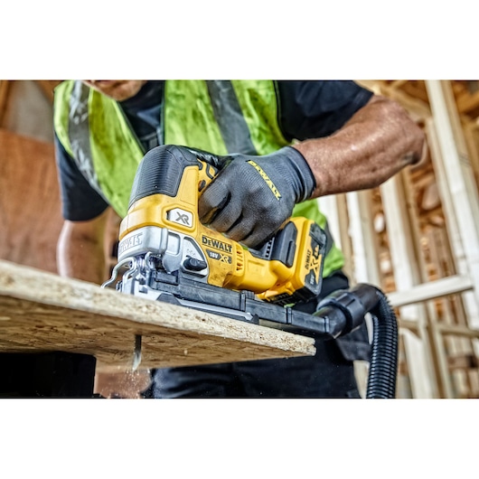A person wearing work gloves and a safety vest uses a DEWALT 18V XR cordless jigsaw to cut through a sheet of wood in a construction setting.