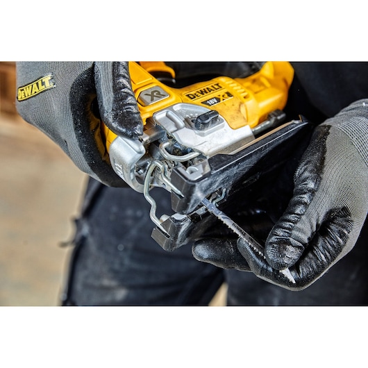 Close-up of a person wearing gloves handling a DEWALT 18V XR jigsaw, preparing to insert a jigsaw blade into the saw.