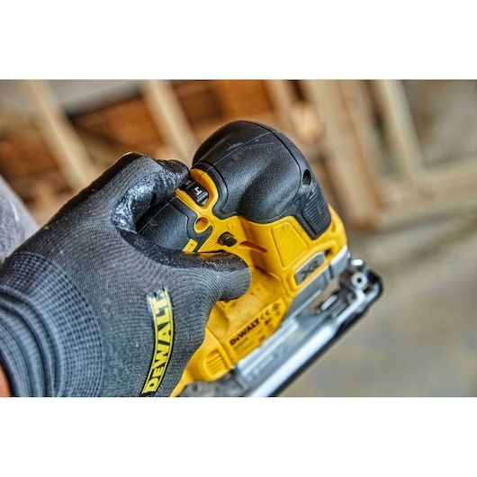 A close-up of a gloved hand adjusting the control on a yellow DEWALT cordless jigsaw, model DCS334P2, with a blurred workshop background.