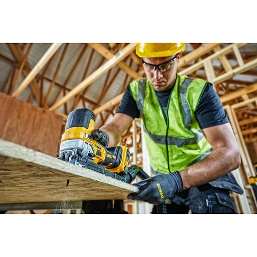 A person wearing a high-visibility vest, gloves, and a hard hat is using a DEWALT DCS335N jigsaw to cut a wooden board at a construction site. The background shows a wooden frame structure.