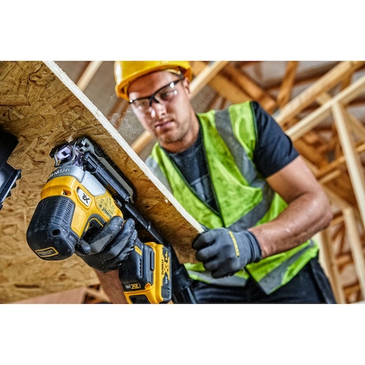 A construction worker wearing a high-visibility vest and gloves is using a DEWALT DCS335N cordless jigsaw to cut a sheet of wood at a building site. The worker's face is blurred for privacy.