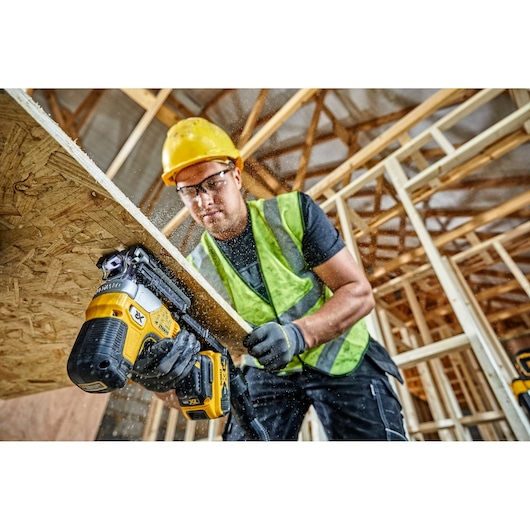 A construction worker wearing a yellow hard hat and safety vest is using a DEWALT DCS335P2 cordless jigsaw to cut a wooden board inside a building under construction. Sawdust is visible around the work area.