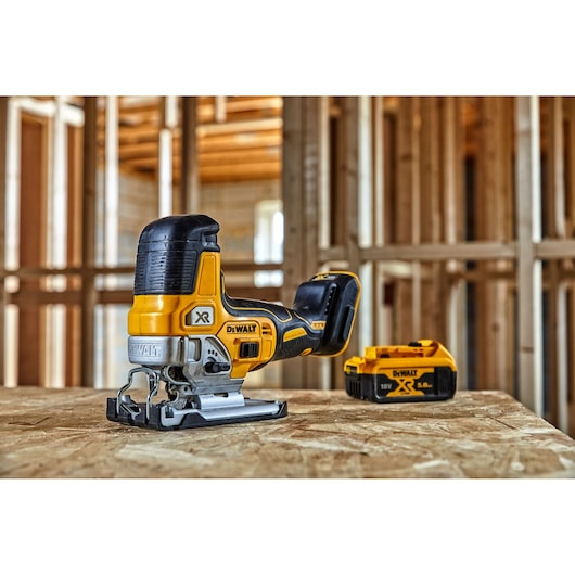 A DEWALT cordless jigsaw and a battery are placed on a wooden surface inside a construction site with exposed wooden framing.