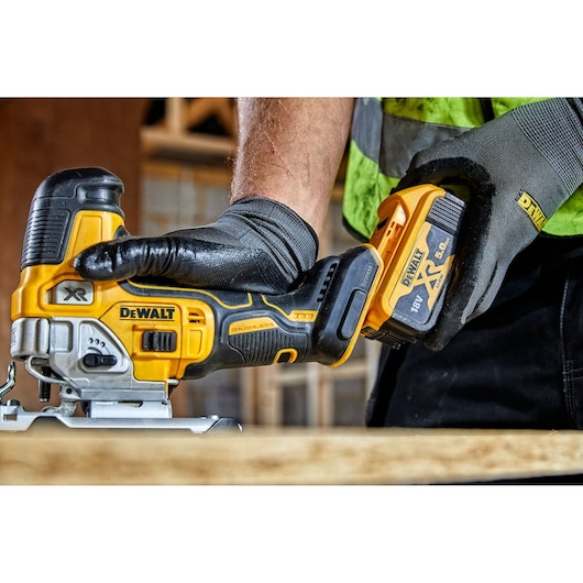 A close-up of a person wearing gloves operating a DEWALT cordless jigsaw, model DCS335P2, with an 18V XR 5.0Ah battery, in a construction setting.