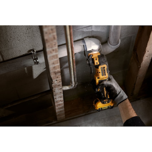 A person wearing a glove is using a yellow DEWALT cordless oscillating multi-tool to cut a pipe against a wall in a