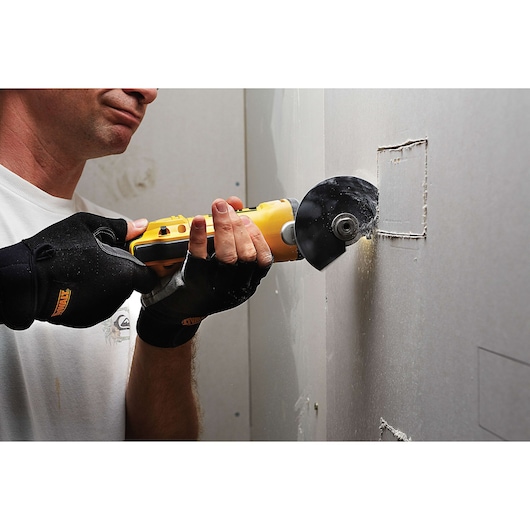 A person wearing protective gloves is using a yellow DEWALT oscillating multi-tool to cut a square section out of a drywall