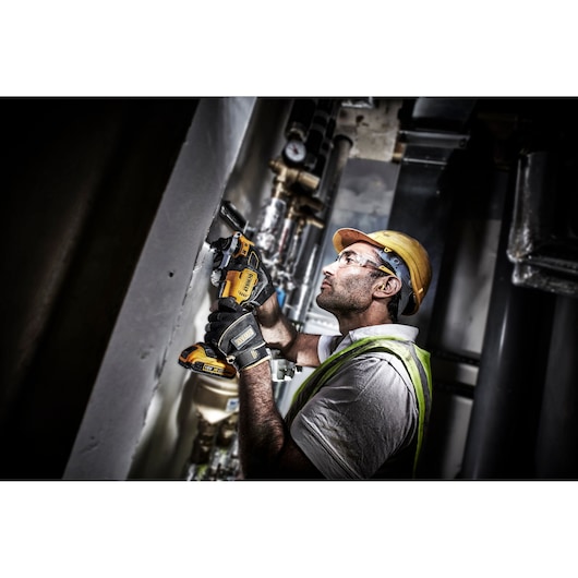 A worker wearing a high-visibility vest, gloves, and a hard hat is using a DEWALT DCS356NT cordless oscillating multi-tool to work on a wall in an industrial environment.