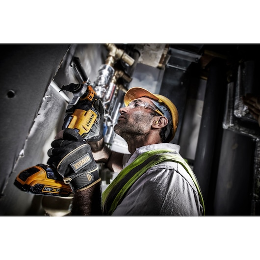 A person wearing a safety helmet, gloves, and a high-visibility vest is using a DEWALT DCS356N cordless oscillating multi-tool to cut into a wall or panel in an industrial or construction setting.