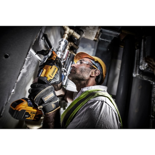A person wearing safety gear operates a DEWALT DCS356P2 cordless oscillating multi-tool near a wall in an industrial environment. The individual is also wearing protective gloves and a high-visibility vest.