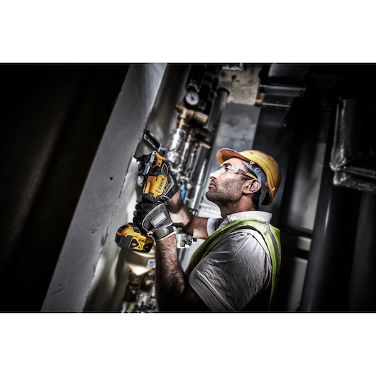 A worker wearing a yellow hard hat and safety vest is using a DEWALT DCS356P2 cordless oscillating multi-tool against a wall in an industrial setting. The worker is also wearing safety gloves and appears to be performing maintenance or installation tasks.