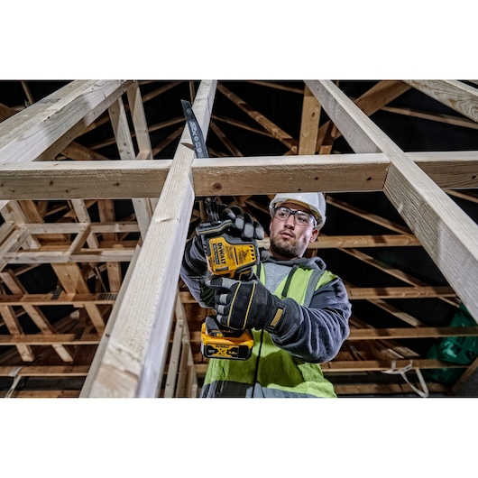 A construction worker wearing gloves and a safety vest uses a DEWALT DCS369P2 cordless reciprocating saw to cut a wooden beam in an unfinished building framework.