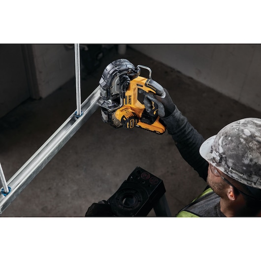A construction worker wearing safety gear uses a yellow DEWALT DCS377NT portable band saw to cut a metal pipe fixed to a steel frame.
