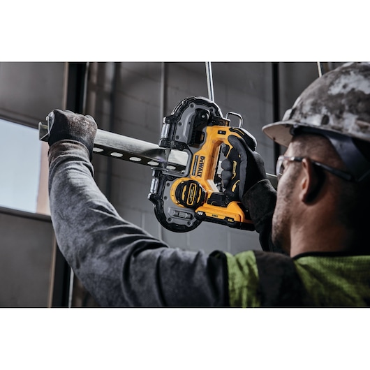 A person wearing gloves and safety gear uses a DEWALT DCS377NT cordless band saw to cut a metal pipe indoors.
