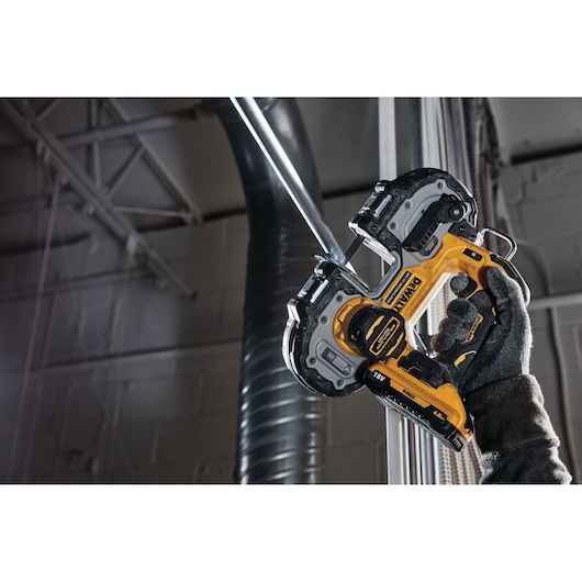A person wearing a dark work glove is using a DEWALT DCS377NT cordless band saw to cut a metal pipe in an industrial setting.