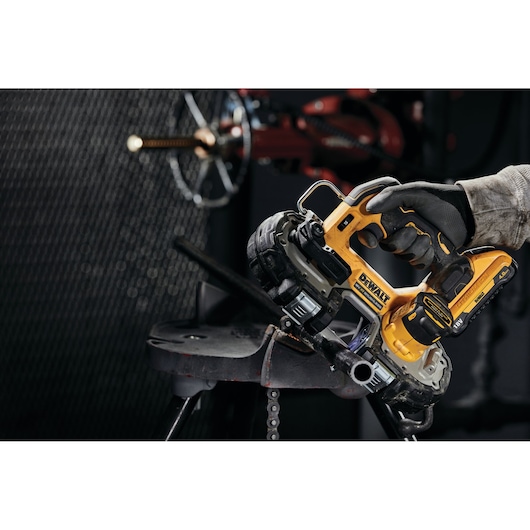 A close-up of a person using a DEWALT cordless bandsaw (model DCS377NT) to cut a metal pipe in an industrial setting. The person's hand is wearing a work glove and is holding the yellow and black power tool.