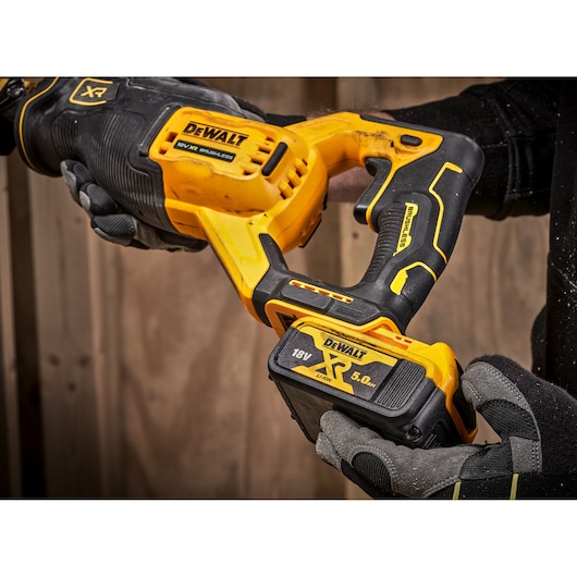 Gloved hands hold a yellow DEWALT 18V XR cordless tool with a visible battery, against a wood background.