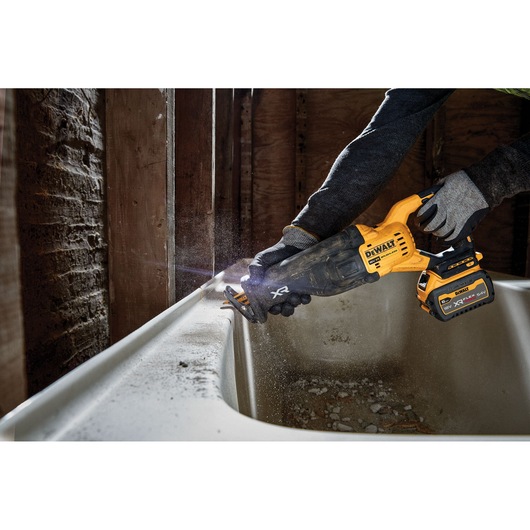 DEWALT cordless reciprocating saw cutting a bathtub edge, operated by gloved hands in a construction setting.