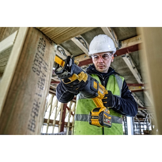 Person uses a Dewalt cordless reciprocating saw on wood at a construction site. Safety gear and gloves are worn.