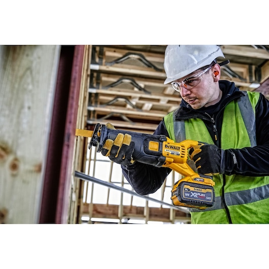 Person using a DEWALT DCS389N cordless reciprocating saw at a construction site with safety gear.