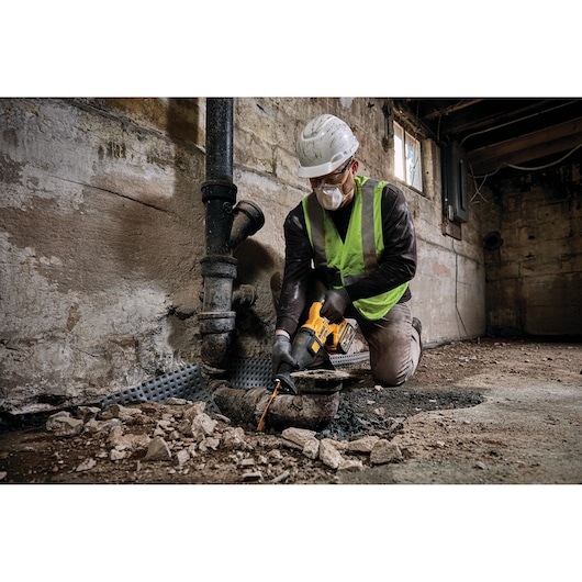 Worker using a DEWALT cordless saw to cut a pipe in a construction area, wearing safety gear and kneeling.