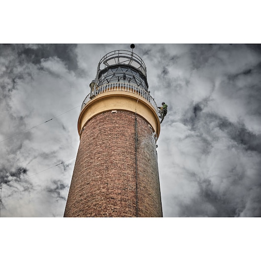 Worker climbs near the top of a tall brick lighthouse against a cloudy sky.