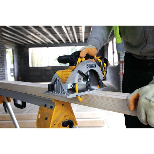 A person wearing work gloves is using a DEWALT cordless circular saw to cut a wooden plank in a construction environment.