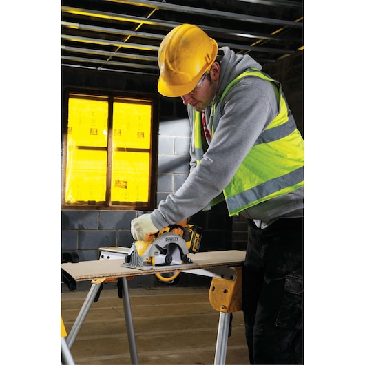 A person wearing a yellow safety helmet and reflective vest is using a DEWALT DCS391NT circular saw to cut wood on a