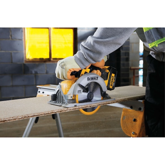 A person wearing protective gloves is using a DEWALT cordless circular saw to cut a wooden board on a sawhorse inside a