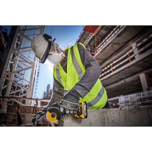 A worker wearing a hard hat, face shield, gloves, and a high-visibility vest uses a DEWALT cordless angle grinder (model DCS438E2T) on a concrete slab at a construction site.