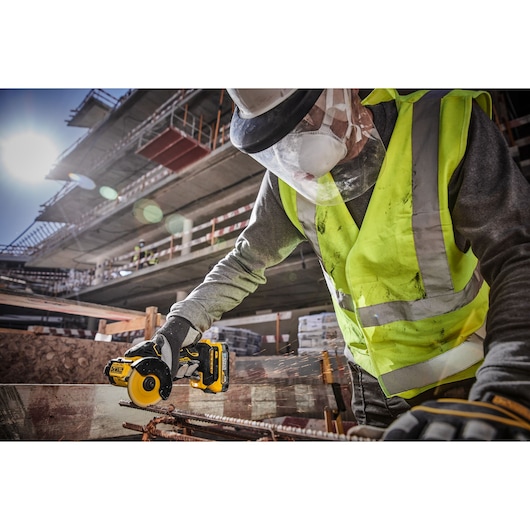 A worker wearing a high-visibility vest, hard hat, face shield, and gloves is using a DEWALT cordless rotary tool to cut metal rebar at a construction site.