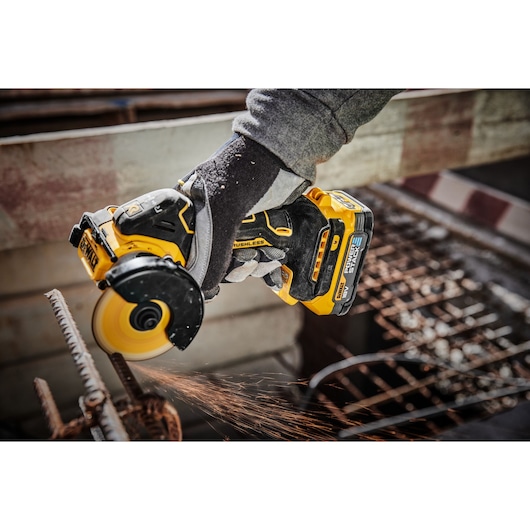 A person wearing work gloves uses a DEWALT cordless brushless angle grinder to cut through a metal rod, with visible sparks flying during the process.