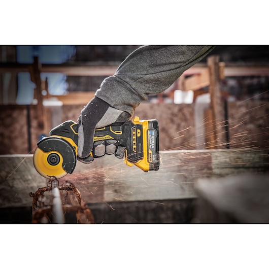 A person wearing gloves operates a DEWALT cordless brushless angle grinder, cutting through a metal rod and producing sparks.