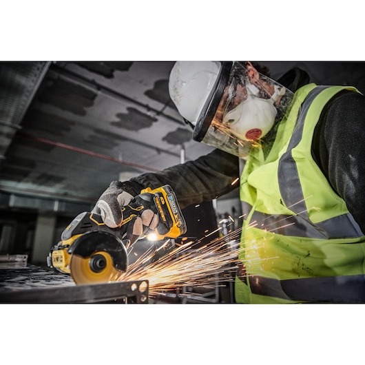 A person wearing safety gear, including a helmet, face shield, mask, gloves, and a high-visibility vest, is using a DEWALT DCS438N cordless power tool to cut metal, creating sparks.