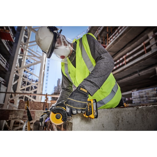 A construction worker wearing a safety helmet, face shield, reflective vest, and gloves is using a DEWALT DCS438N cordless tool to cut through concrete at an outdoor construction site.