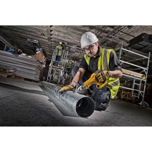 Worker uses DEWALT cordless metal shear on duct pipe at construction site. Safety gear and tools visible.